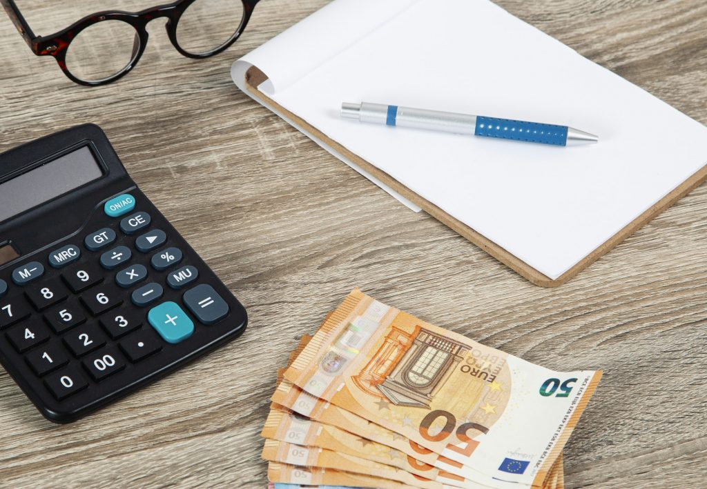 Ein Taschenrechner, ein 50-Euro-Schein und ein Stift auf einem Tisch, symbolisch für Buchaltungsdienstleistung, leiten den Nutzer zur 'Über Uns' Seite von Accounting Solutions.